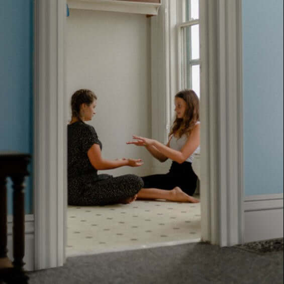 Two women seated on the floor in a room, engaged in a hands-on activity, with natural light coming through the windows.